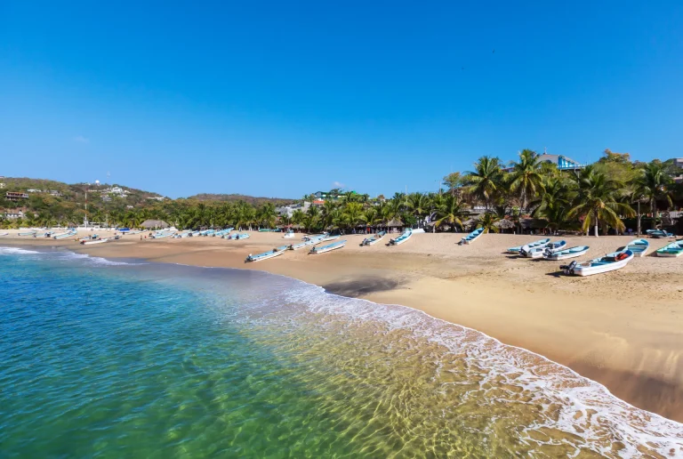 Playa de arena dorada con aguas turquesa, palmeras y lanchas en la costa del Pacífico mexicano, similar a las bahías de Huatulco, Oaxaca